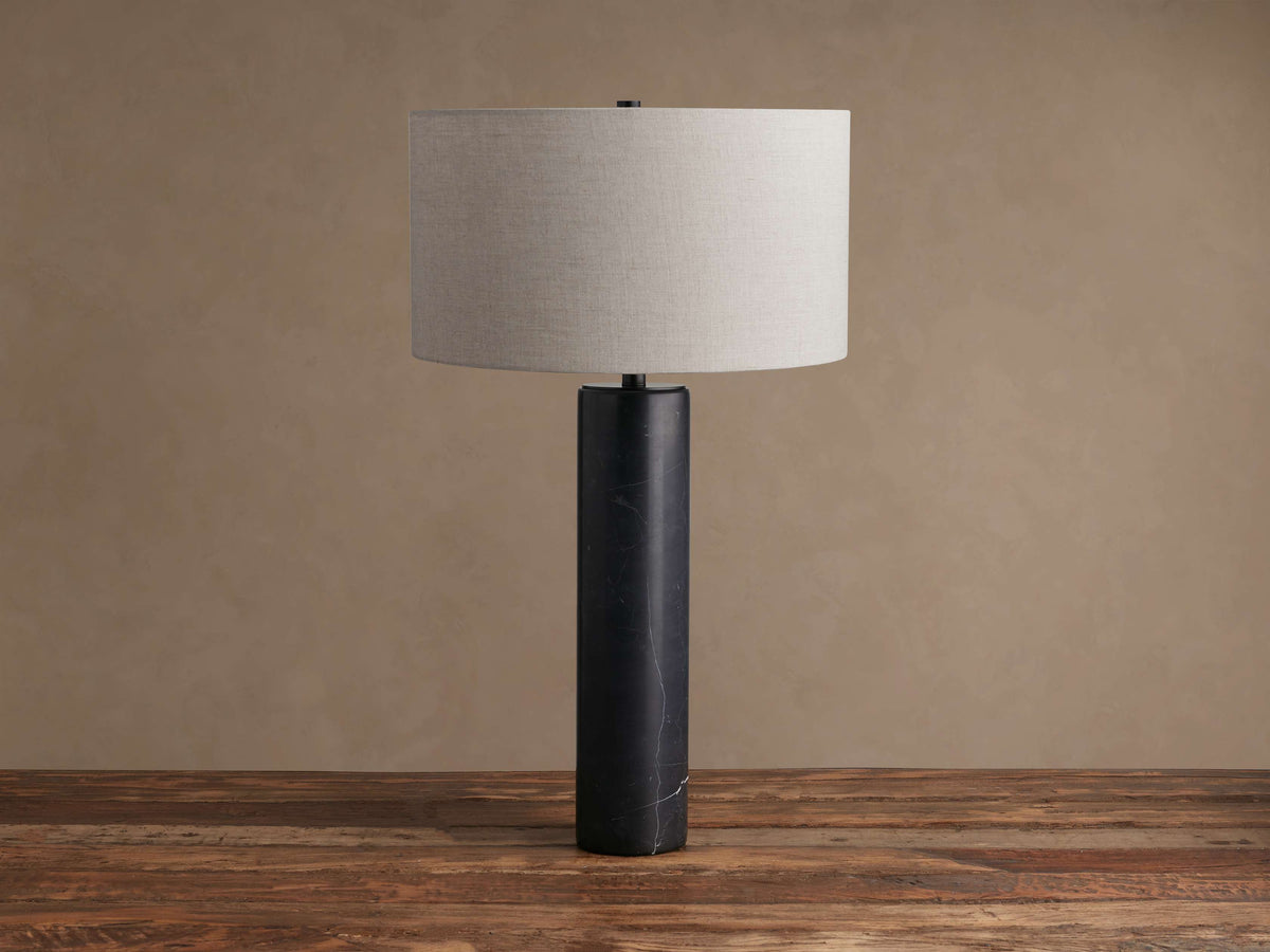 Close-up of Brixton Table Lamp. Its cylindrical dark marble base with white veining meets a black metal stem on a beige background.