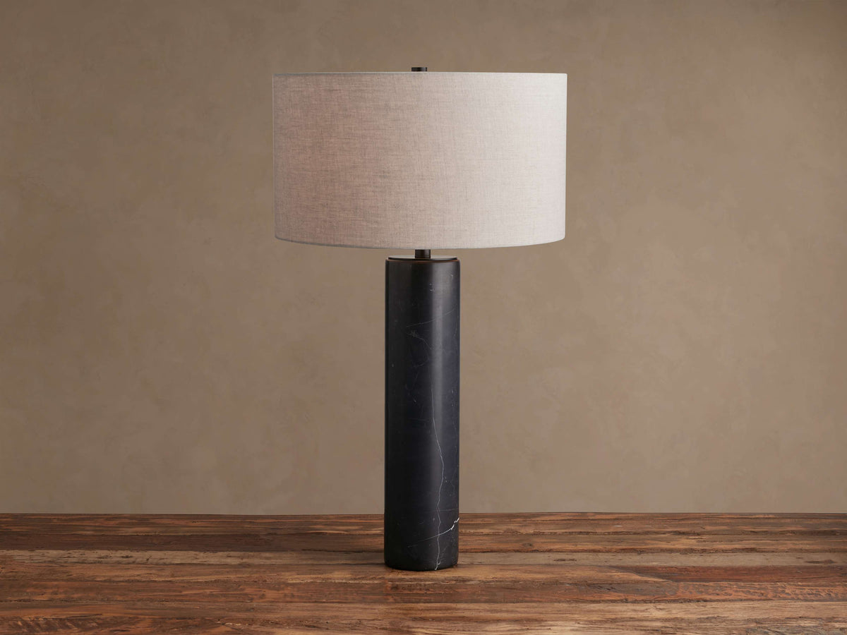 Brixton Table Lamp with a black marble cylindrical base and a light gray fabric drum shade, standing on a wooden surface.