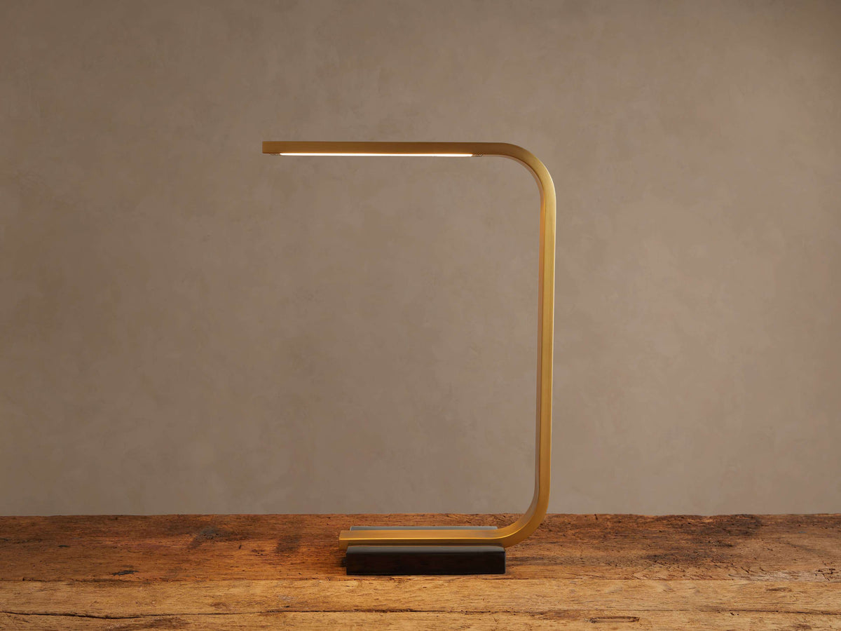 Karson Task Table Lamp: sleek brass, U-shaped lamp with an illuminated LED strip, on a black marble base, resting on a wooden table.