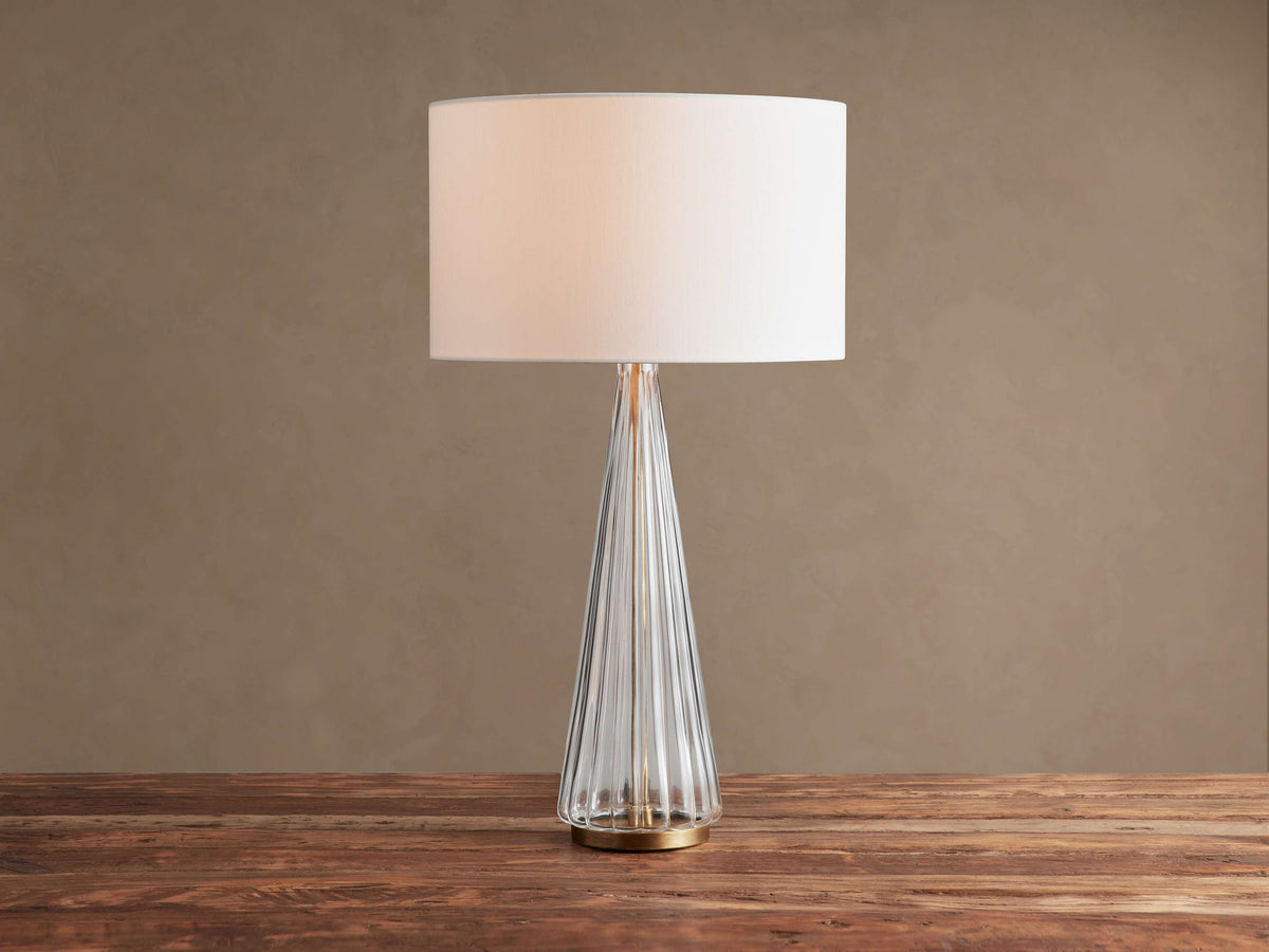 Elisabetta Table Lamp: white linen shade, clear fluted glass conical base, brass finish. On a rustic wood table.