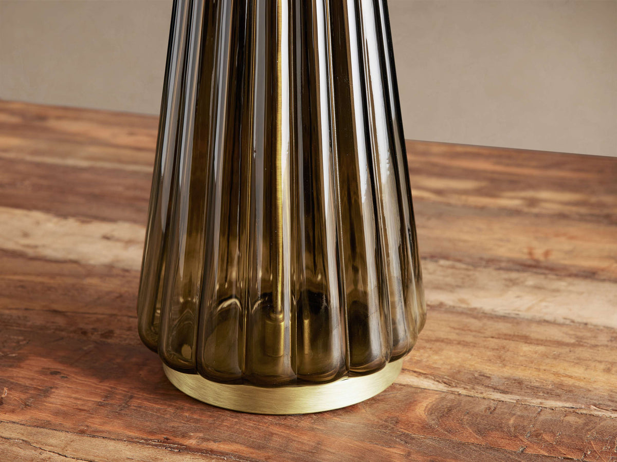 Elisabetta Table Lamp, close-up of its fluted smoky glass base and round brass-finished steel base on a wooden table.