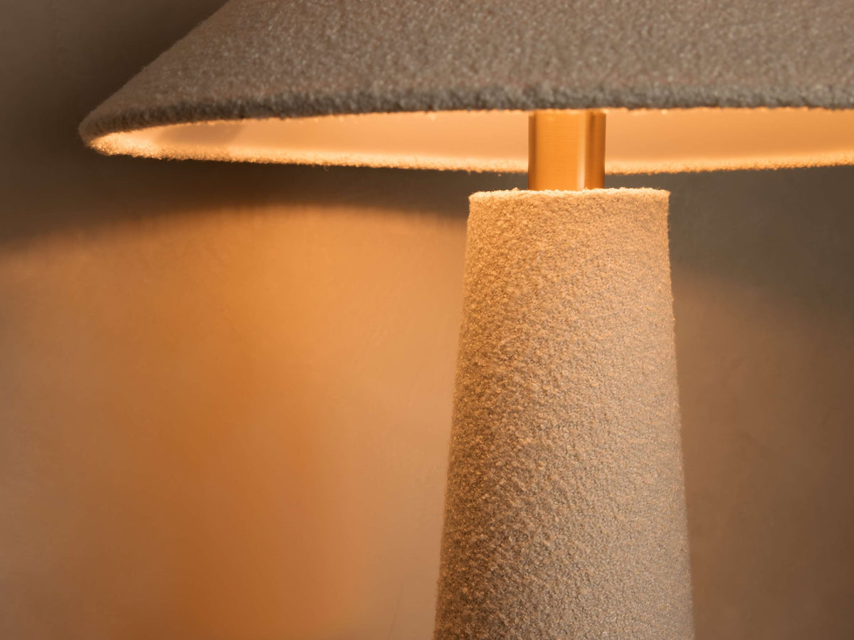 Close-up of textured conical lamp base in light beige. Textured gray conical shade above a golden rod, casting warm light on wall.