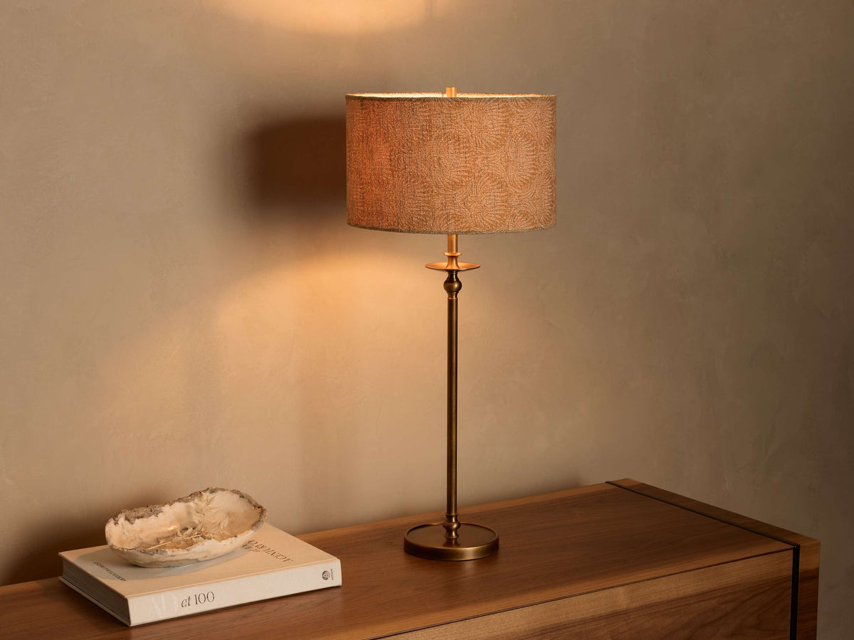 Front view of the Winslow Table Lamp with an antique brass finish base and a light brown patterned fabric shade, on a wooden table.