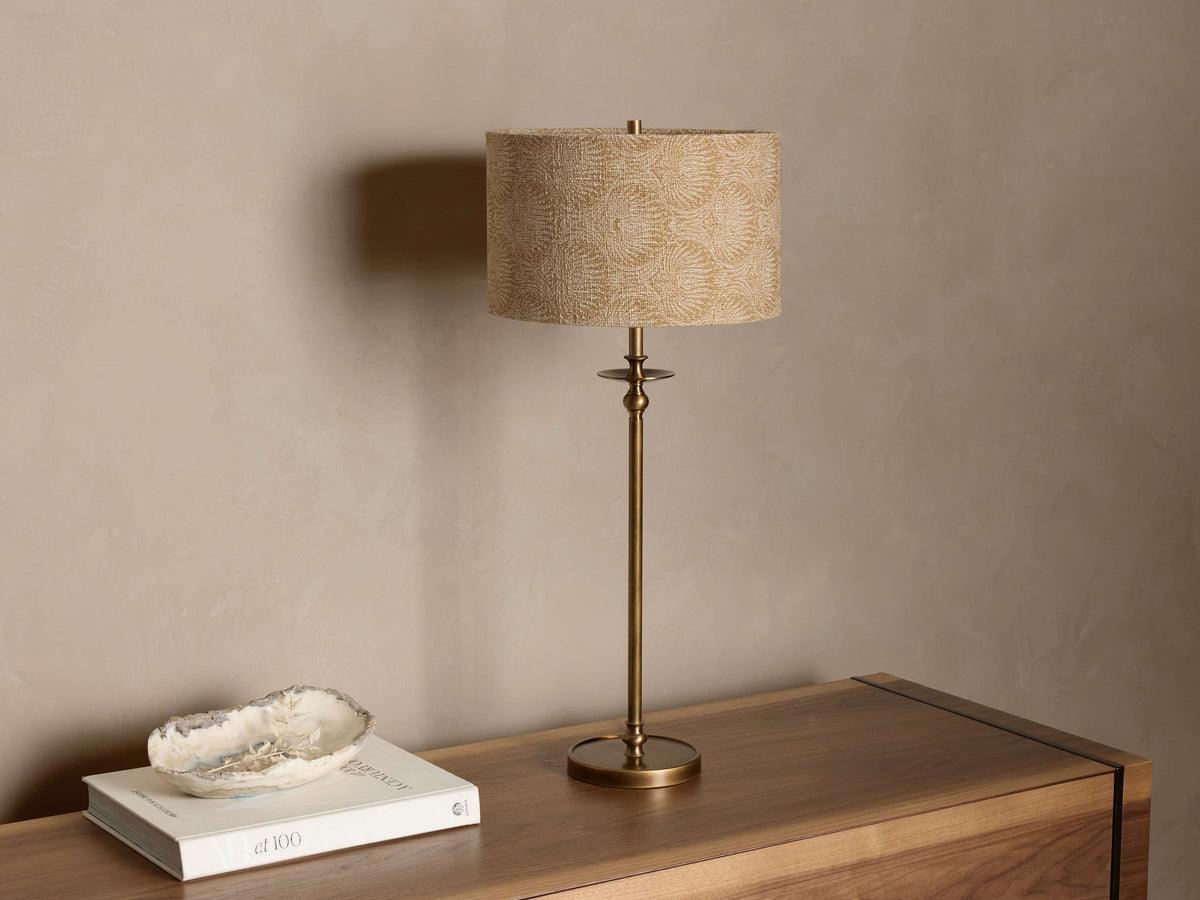 Front view of the antique brass Winslow Table Lamp with a light beige patterned fabric shade on a wooden table.