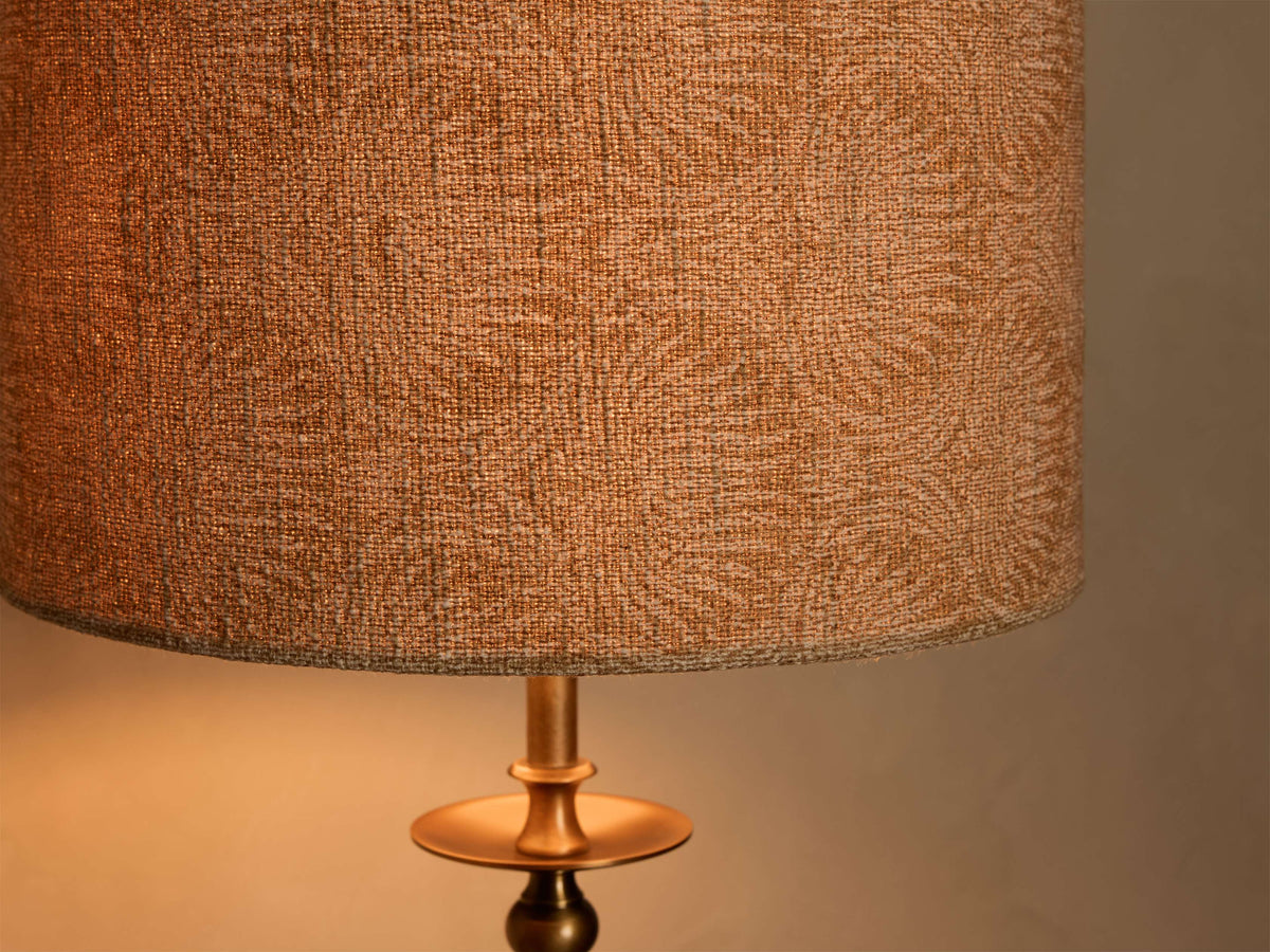 Close-up of the lit Winslow Table Lamp, highlighting its textured tan fabric shade and antique brass base with a warm glow.