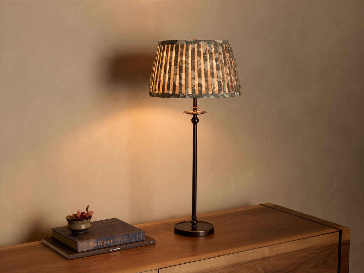 Illuminated Winslow Table Lamp with a bronze-finish iron base and a light brown patterned fabric drum shade on a wooden surface.
