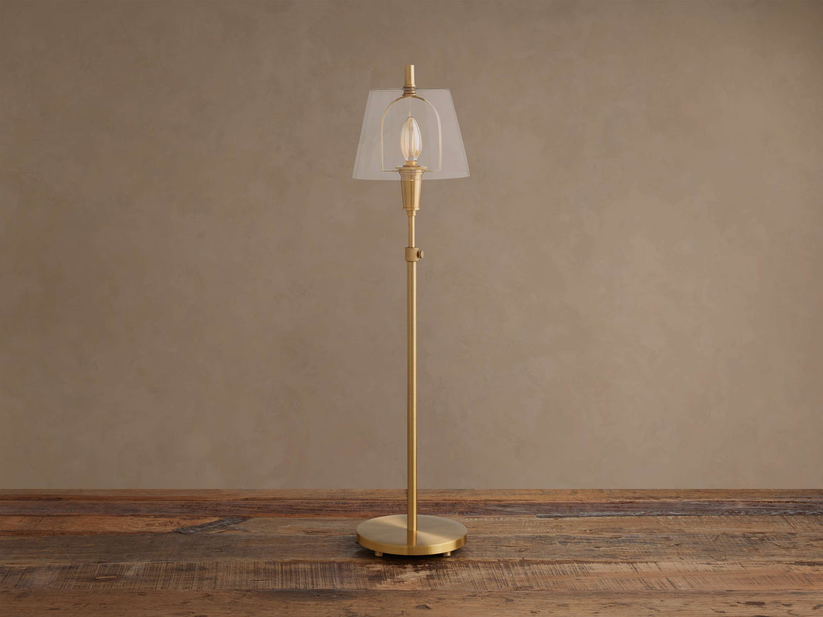 Brass Candice Table Lamp with a clear conical shade and illuminated bulb, on a wooden surface against a textured beige wall.