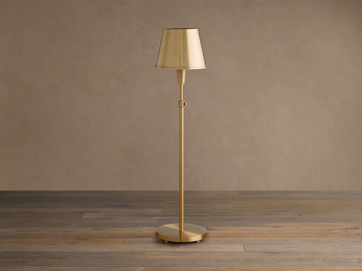 Candice Table Lamp. Tall, slender brass lamp with a tapered brass shade, slim stem, and round base, standing on a wooden floor.
