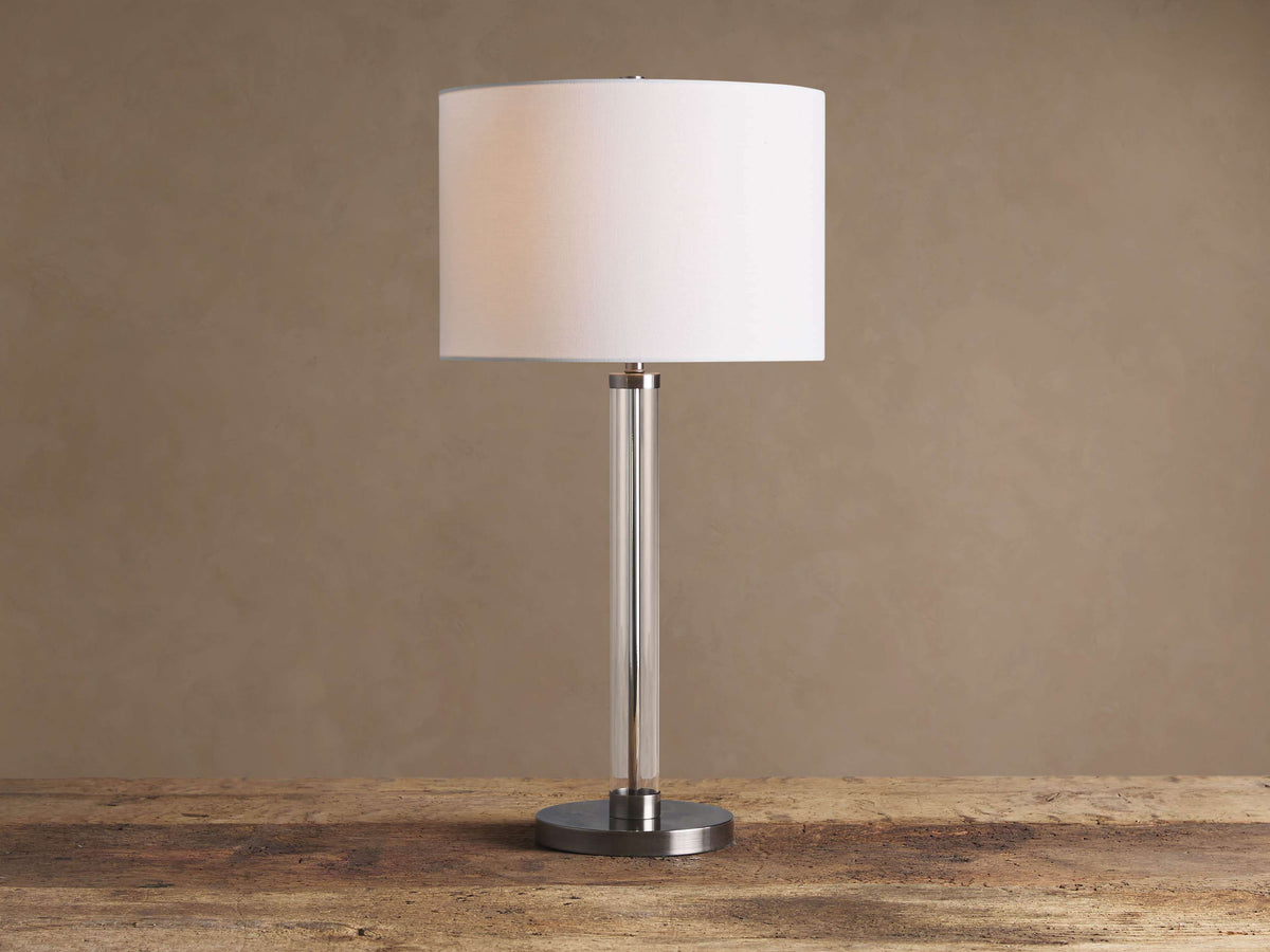 Glass Column Table Lamp with clear glass column, brushed metal base, and white linen shade on a wooden table.