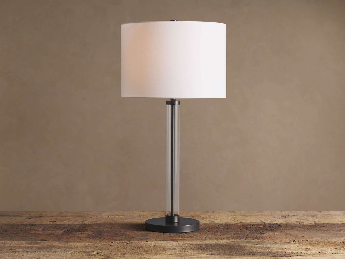 Glass Column Table Lamp with clear glass column, dark iron base, and white linen drum shade on a rustic wood table.