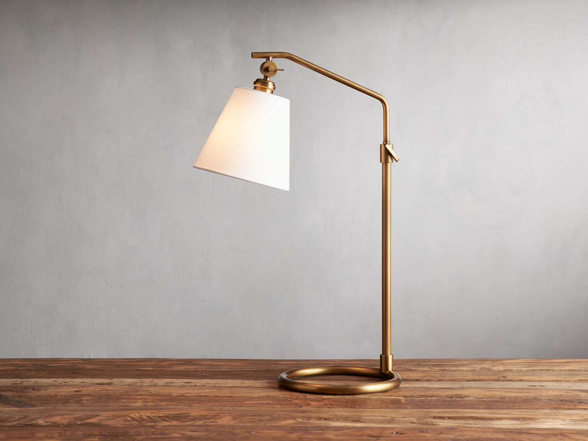 Kellen Table Lamp in antiqued-brass with white linen shade, on wood surface. Adjustable arm and round base, cord visible.