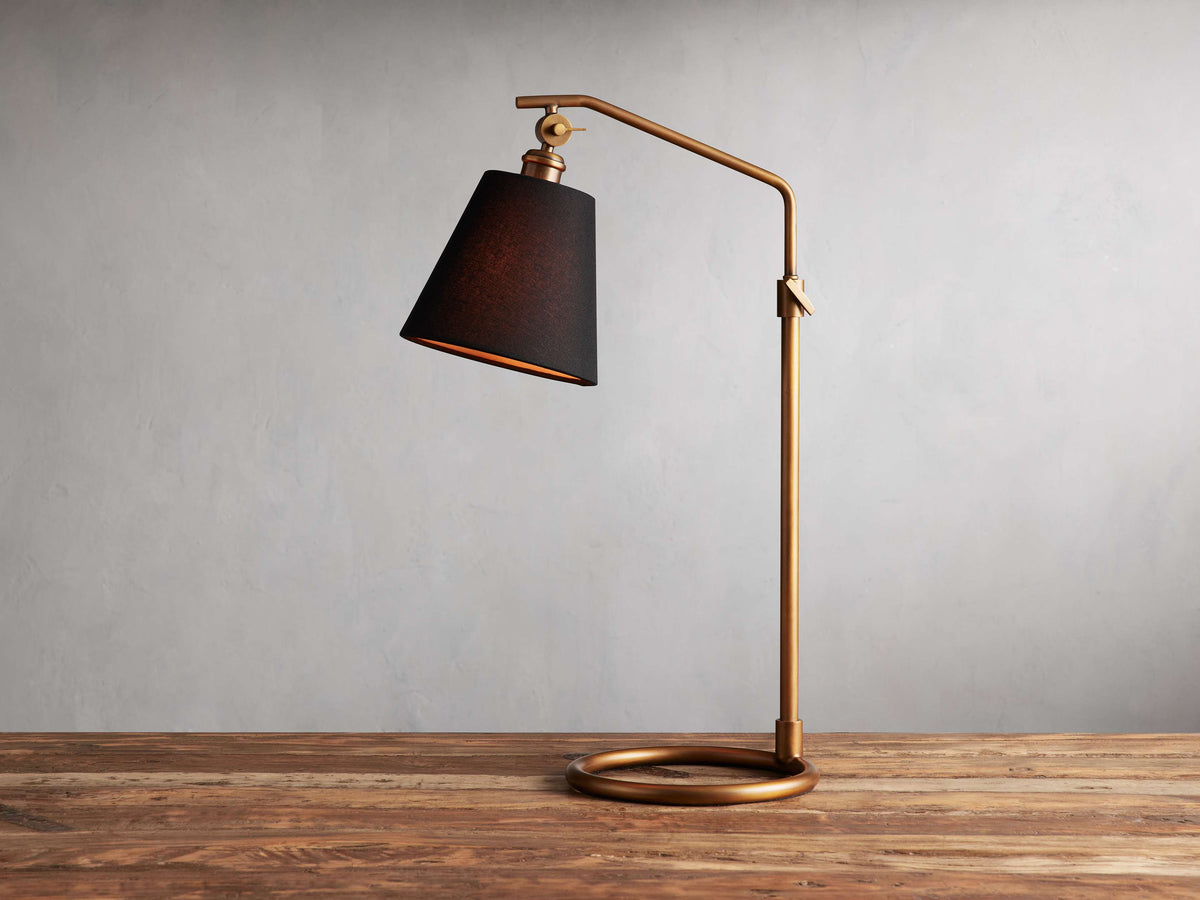 Kellen Antiqued Brass Table Lamp with dark tapered linen shade on adjustable brass stand, placed on wooden surface.