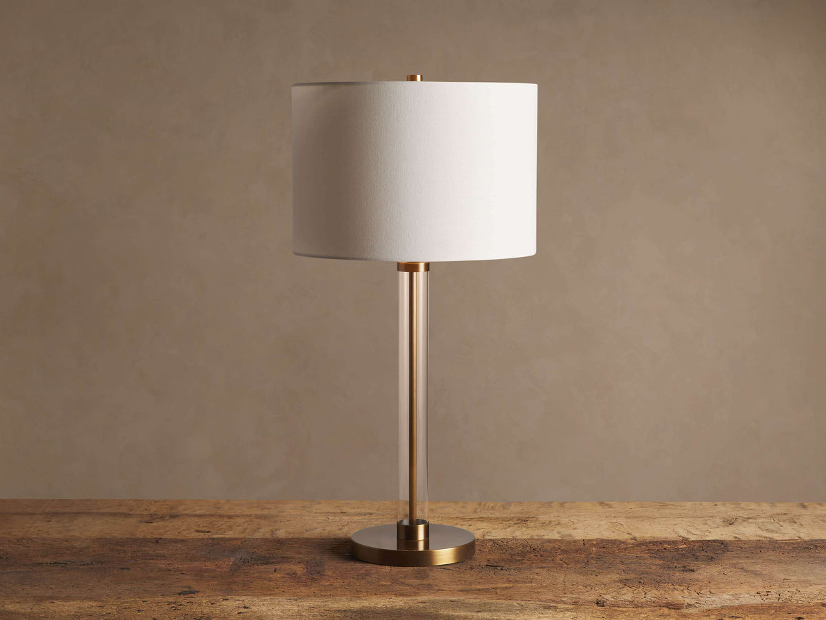 Glass Column Table Lamp with clear glass column, white linen shade, and gold base on a rustic wood table.