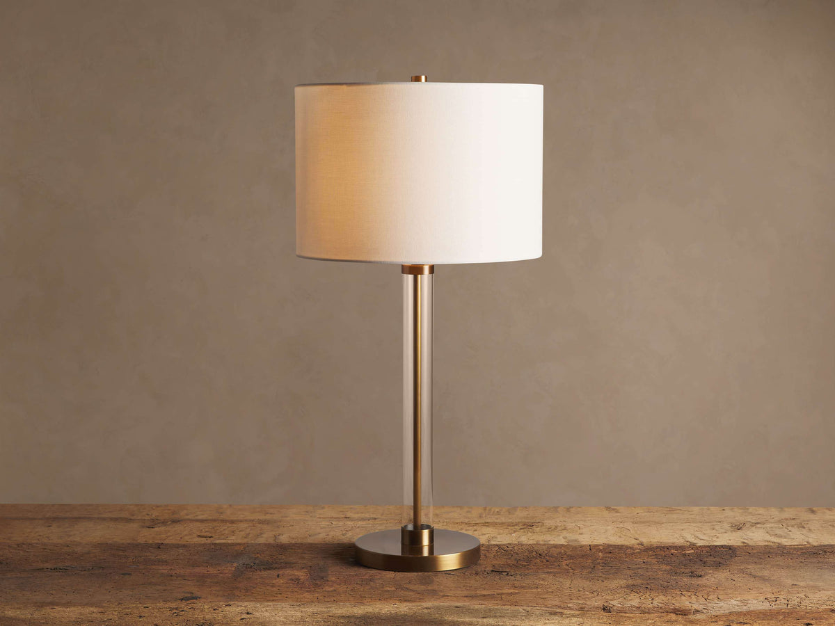 Lit Glass Column Table Lamp featuring a clear glass stem, bronze base, and cream linen drum shade on a rustic wood table.