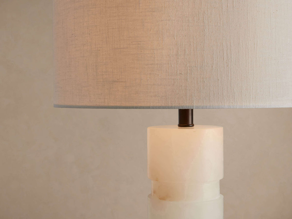 Close-up of lit Novalie Table Lamp: translucent alabaster base, bronze accent, and cylindrical light-colored fabric shade.
