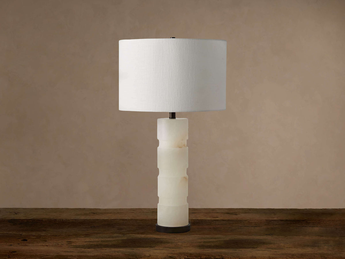 Novalie Table Lamp with a white cylindrical shade, carved alabaster base, and dark bronze bottom, on a wooden table.