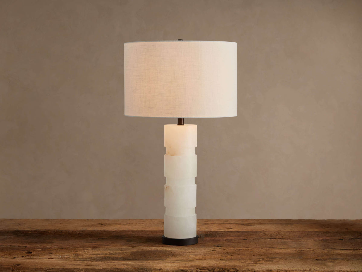 Novalie Table Lamp: Carved white alabaster body with bronze base, topped by a large off-white fabric drum shade on a wooden surface.