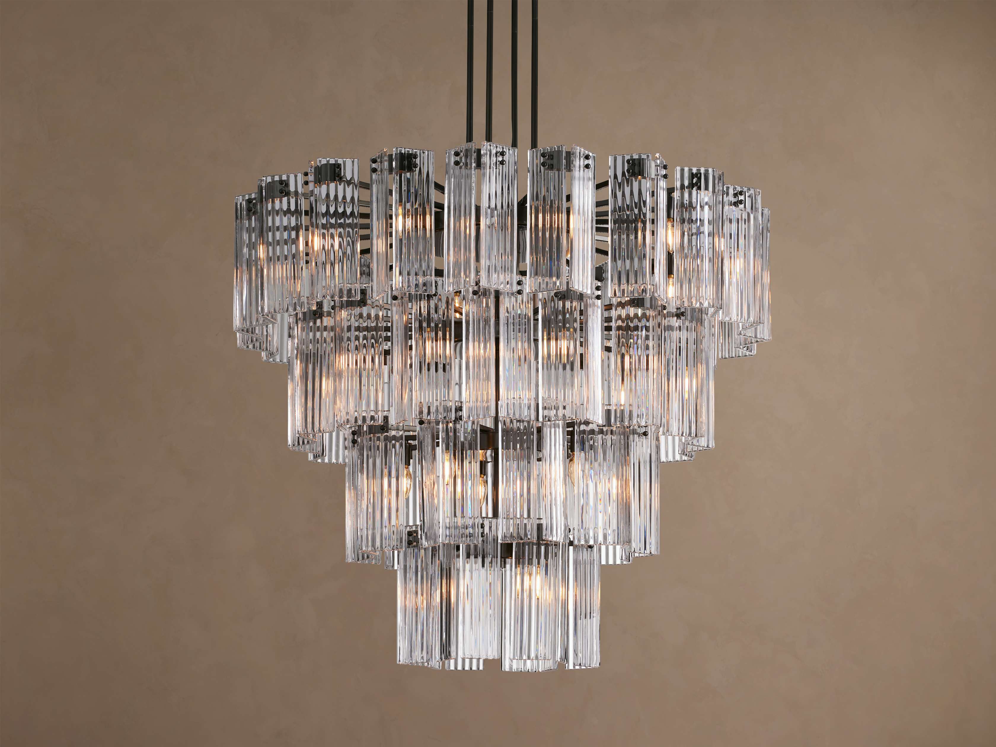 Delsie Round Chandelier with illuminated tiers of clear, fluted glass rods and a dark metal frame hanging against a taupe wall.