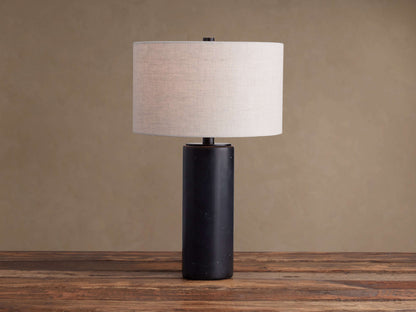 Brixton Table Lamp with a dark, cylindrical marble base & light fabric drum shade on a rustic wooden table.