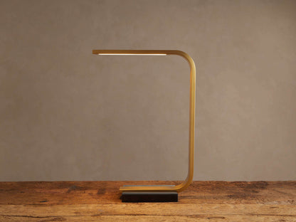 Karson Task Table Lamp: sleek brass, U-shaped lamp with an illuminated LED strip, on a black marble base, resting on a wooden table.