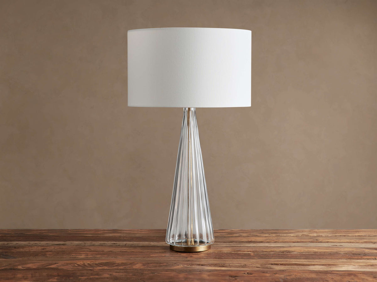 Elisabetta Table Lamp with a clear fluted glass tapered base, brass bottom, and white linen shade on a rustic wood table.