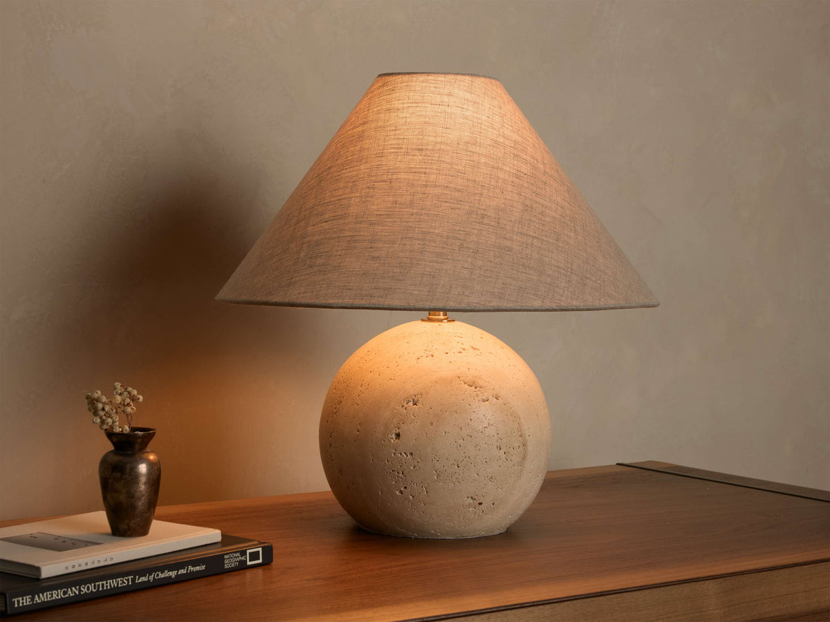 Front view of a table lamp featuring a conical gray linen shade and a spherical, porous beige stone base on a wood surface.