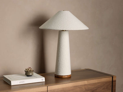 Cream textured table lamp with conical shade, base, brass connector, and wooden foot, on a wooden surface.