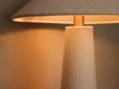 Close-up of textured conical lamp base in light beige. Textured gray conical shade above a golden rod, casting warm light on wall.