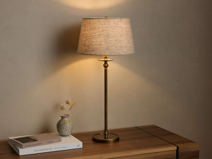 Front view of the Winslow Table Lamp with an antique brass base and light grey fabric shade, illuminated, on a wooden table.