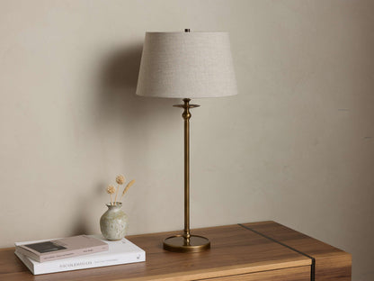Front view of the antique brass Winslow Table Lamp with a natural linen-colored tapered drum shade, on a wooden table.