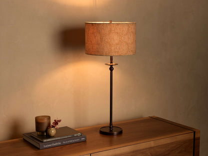 Illuminated Winslow Table Lamp with a bronze-finish iron base and a light brown patterned fabric drum shade on a wooden surface.