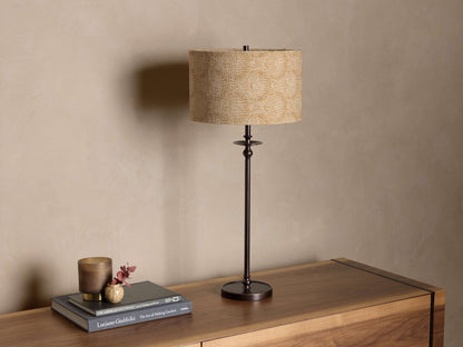 Front view of the Winslow Table Lamp with an antique bronze finish base and a patterned beige fabric shade on a wooden credenza.