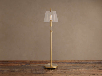 Brass Candice Table Lamp with a clear conical shade and illuminated bulb, on a wooden surface against a textured beige wall.