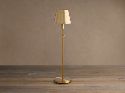 Candice Table Lamp. Tall, slender brass lamp with a tapered brass shade, slim stem, and round base, standing on a wooden floor.