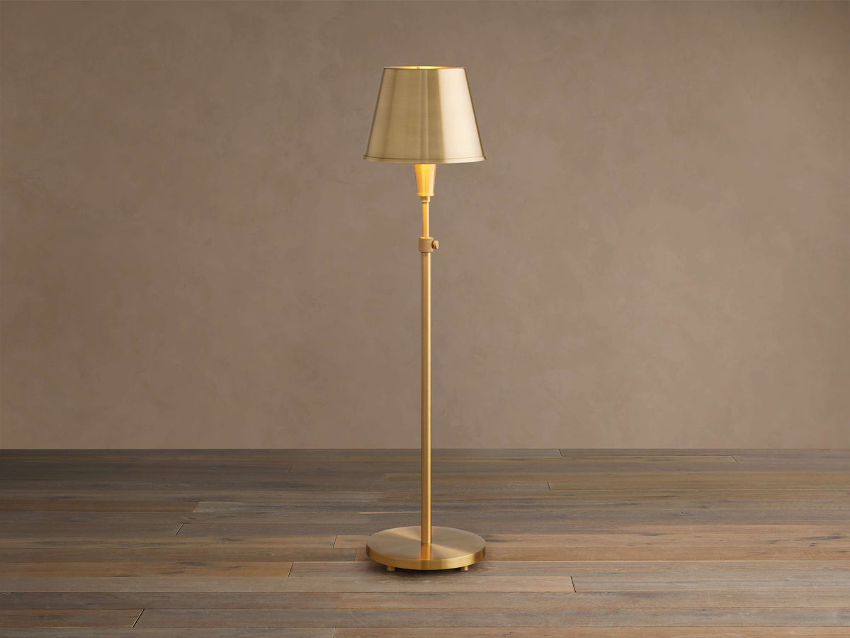Illuminated Candice Table Lamp in brass, with a cone-shaped brass shade, standing on a wooden floor against a beige wall.