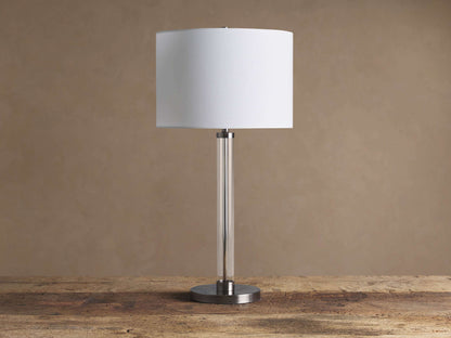 Glass Column Table Lamp: clear glass column, white linen shade, and round dark iron base, on a rustic wooden table.