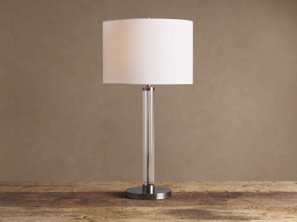 Glass Column Table Lamp with clear glass column, brushed metal base, and white linen shade on a wooden table.