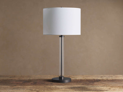 A Glass Column Table Lamp with white cylindrical shade, clear glass column, and dark metal base on a wooden table.
