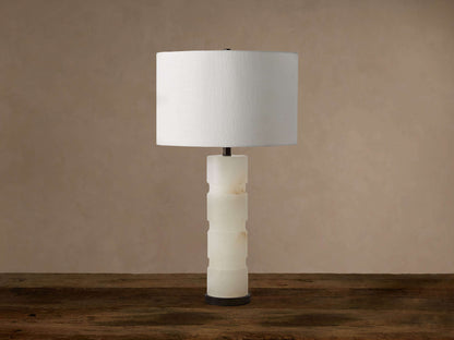 Novalie Table Lamp with a white cylindrical shade, carved alabaster base, and dark bronze bottom, on a wooden table.