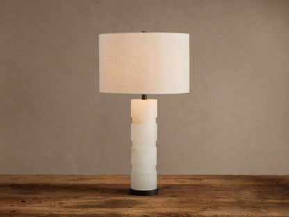 Novalie Table Lamp: Carved white alabaster body with bronze base, topped by a large off-white fabric drum shade on a wooden surface.