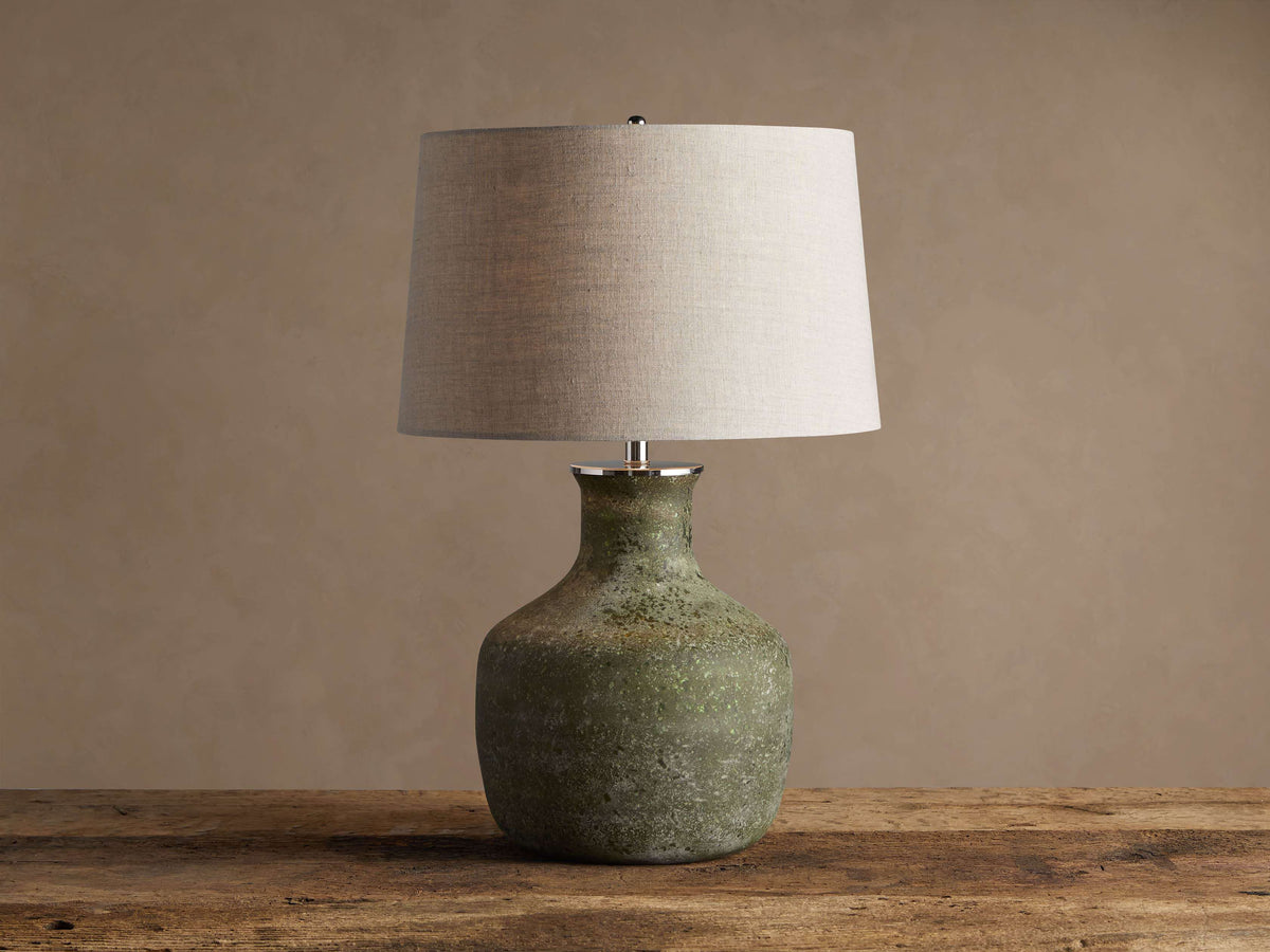 Galena Table Lamp: Green, sand-blasted glass bottle-shaped base with a light linen shade on a rustic wood surface.