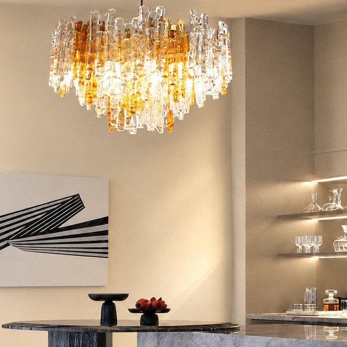 A modern multi-tiered crystal chandelier with amber glass accents hangs over a dining table in a contemporary kitchen and bar setting.