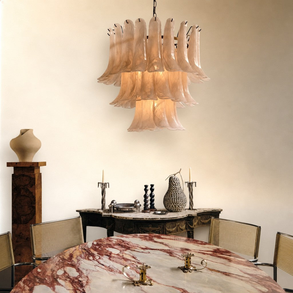 A luxury chandelier with multiple tiers of fluted, bell-shaped glass shades in a warm blush pink hue, hanging in a styled dining room above a marble table.