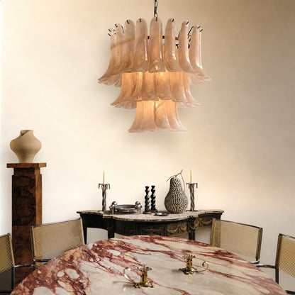 A luxury chandelier with multiple tiers of fluted, bell-shaped glass shades in a warm blush pink hue, hanging in a styled dining room above a marble table.